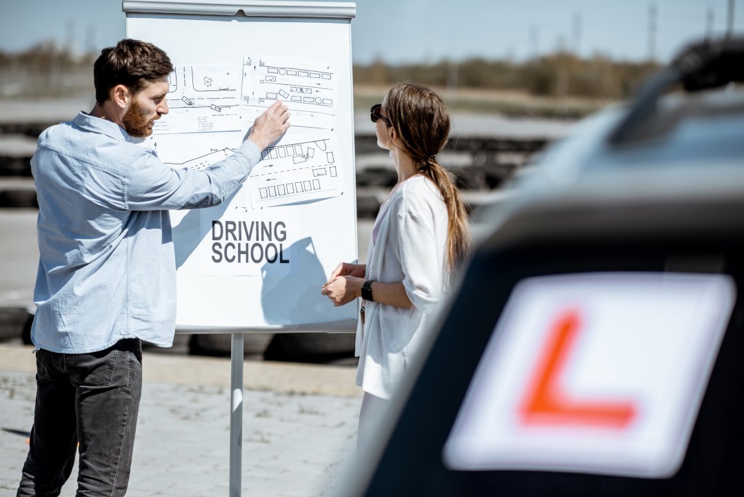 Male private driving lessons instructor showing traffic schemes to a young female student standing on the training ground at the driver's school